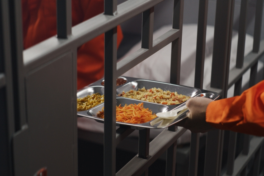 Elderly prisoner in orange uniform sits in prison cell. Prison guard gives him tray of food through metal bars. Guilty criminal serves imprisonment term for crime. Inmate in jail or detention center.